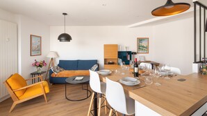 A modern living room with a wooden dining table, white chairs, a blue sofa, and a yellow armchair. - La Nacre - Vue Mer - Two Bedroom Apartment, Sleeps 4 (Erquy)