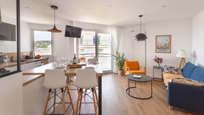 A modern living room with a sofa, a dining table with chairs, a TV, and a balcony with a view. - La Nacre - Vue Mer - Two Bedroom Apartment, Sleeps 4 (Erquy)
