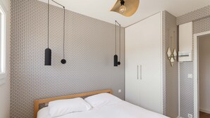 A neatly made bed in a room with patterned wallpaper, a wooden headboard, and a hanging light fixture. - La Nacre - Vue Mer - Two Bedroom Apartment, Sleeps 4 (Erquy)