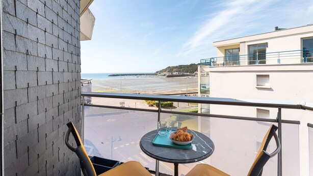A balcony with a table set for two, overlooking a beach and a white building. - La Nacre - Vue Mer - Two Bedroom Apartment, Sleeps 4 (Erquy)