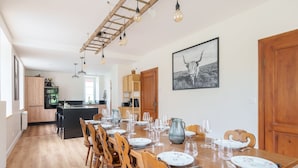 Un comedor con una larga mesa de madera dispuesta para seis personas, rodeada de sillas de madera. Se observa una cocina al fondo con un armario de madera y un refrigerador.