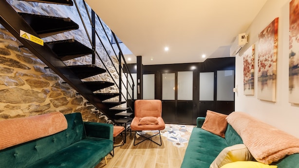 Una sala moderna con pared de piedra, una escalera, un sofá color turquesa, un sillón marrón y una alfombra estampada.