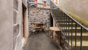 Un estrecho callejón con un muro de piedra, una mesa y sillas de madera, y una escalera de metal.