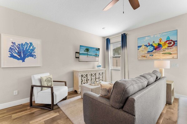 A living room with a grey sofa, a white armchair, a wooden cabinet, and a painting of a beach scene.