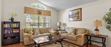 A living room with a sofa, coffee table, and lamps. There is a bookshelf with books and a potted plant.
