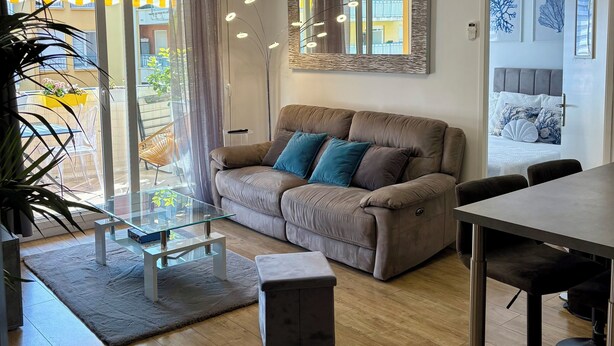 A modern living room with a brown leather sofa, a glass coffee table, a dining table with chairs, and a large mirror on the wall.