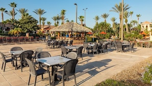 An outdoor patio area with tables and chairs, surrounded by palm trees and a thatched-roof structure. - Rendezvous at Regal Palms - Nearby Theme Parks! (Davenport)