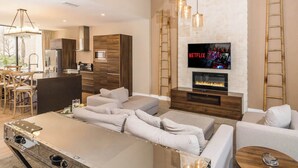 A modern living room with a fireplace, a TV mounted on a white brick wall, and a dining area with wooden furniture.