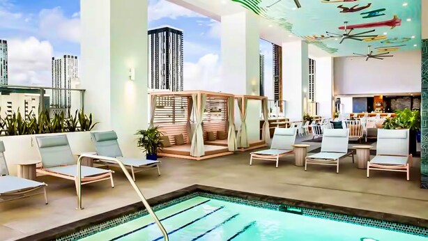 Un espace piscine intérieur moderne, doté de chaises longues, d’un coin salon et offrant une vue sur de hauts immeubles.