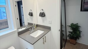 Ett modernt badrum med dubbelt handfat, en sminkbord och en stor spegel. Det finns ett fönster som släpper in naturligt ljus, en toalett och en krukväxt.