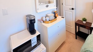 Ett kompakt rum med en vit minibar, en kaffebryggare, ett litet bord med en växt och en dörr som leder till ett annat rum.