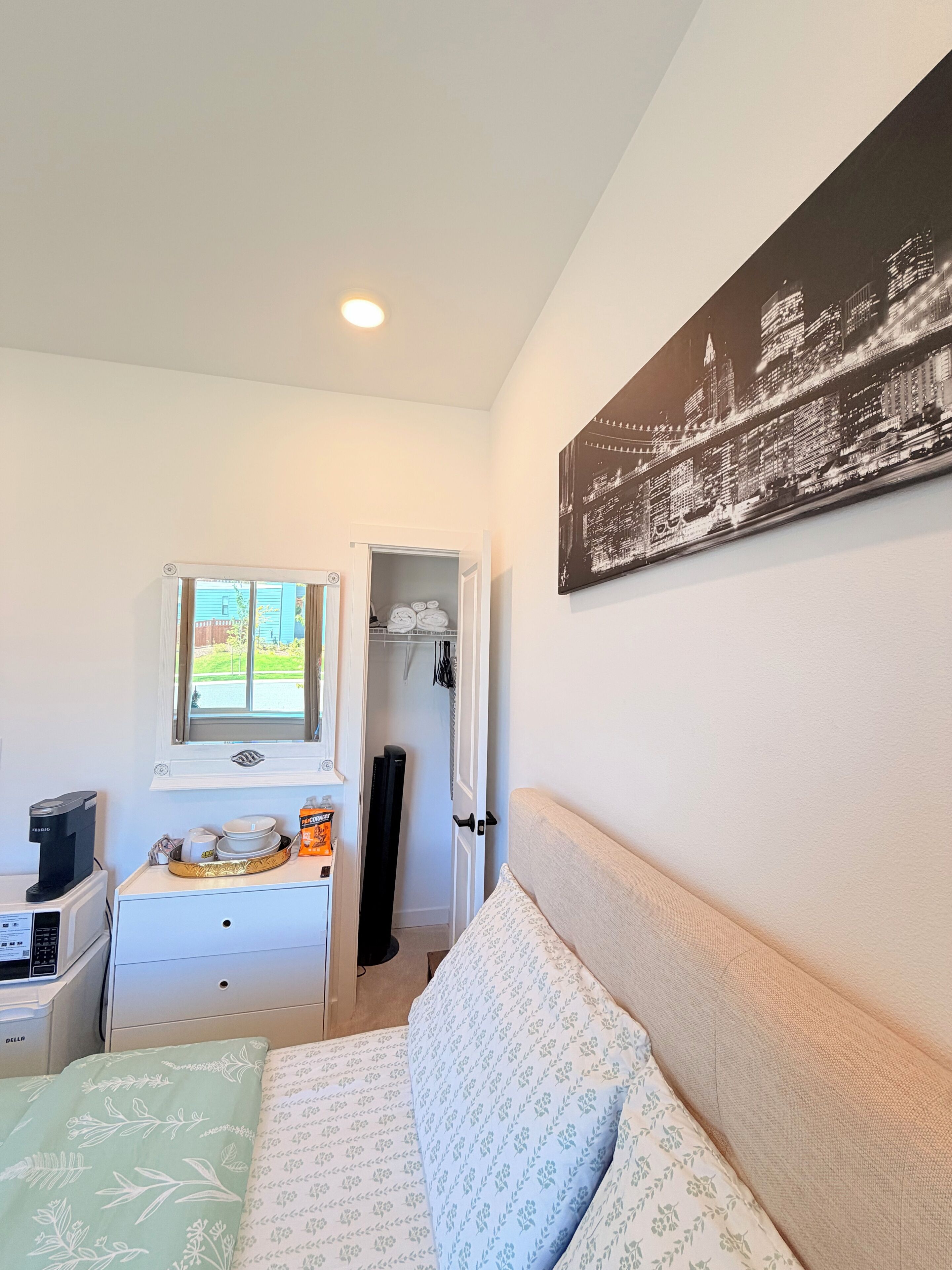 A hotel room with a bed, a desk with a coffee maker, a window with a view, and a cityscape painting on the wall.