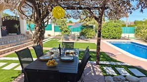 A patio with a table set for dining, a pool, and a garden with a pergola.