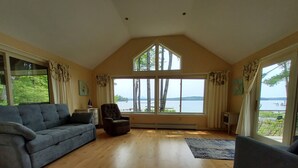A spacious living room with a high ceiling, large windows offering a view of a lake, a blue sofa, a dark armchair, and a small table with a lamp.