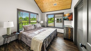 A bedroom with a bed, two bedside lamps, a TV, and a view of a green landscape through large windows.