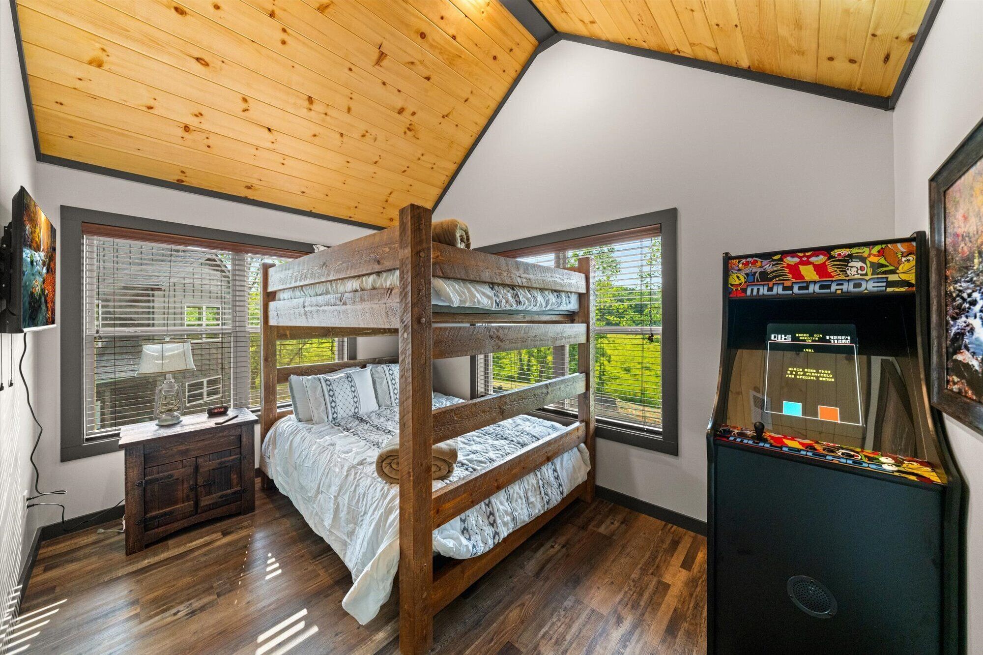 Habitación con literas, techo de madera, ventanas amplias, un mueble pequeño y una consola de videojuegos.
