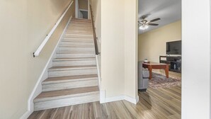 A staircase with wooden steps and white handrails, leading to a room with a ceiling fan, a flat-screen TV, and a kitchen area. - Clean & Cozy 2, Near MLB Airport, HRMC, Food, Nightlife, Beaches & Shopping! (Melbourne)
