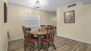 A dining area with a round wooden table and six chairs, a window with blinds, a ceiling light fixture, and a framed picture on the wall. - Clean & Cozy 2, Near MLB Airport, HRMC, Food, Nightlife, Beaches & Shopping! (Melbourne)