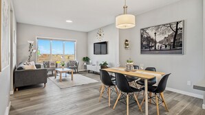 A modern living room with a dining area, a large window, a flat-screen TV, and a cityscape painting. - Cozy 3 Bedroom Home in Rangeview (Calgary)