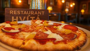 Una pizza con diferentes ingredientes sobre un plato, un letrero que dice "RESTAURANT HVITA" al fondo, y un interior desenfocado que sugiere un espacio para comer.