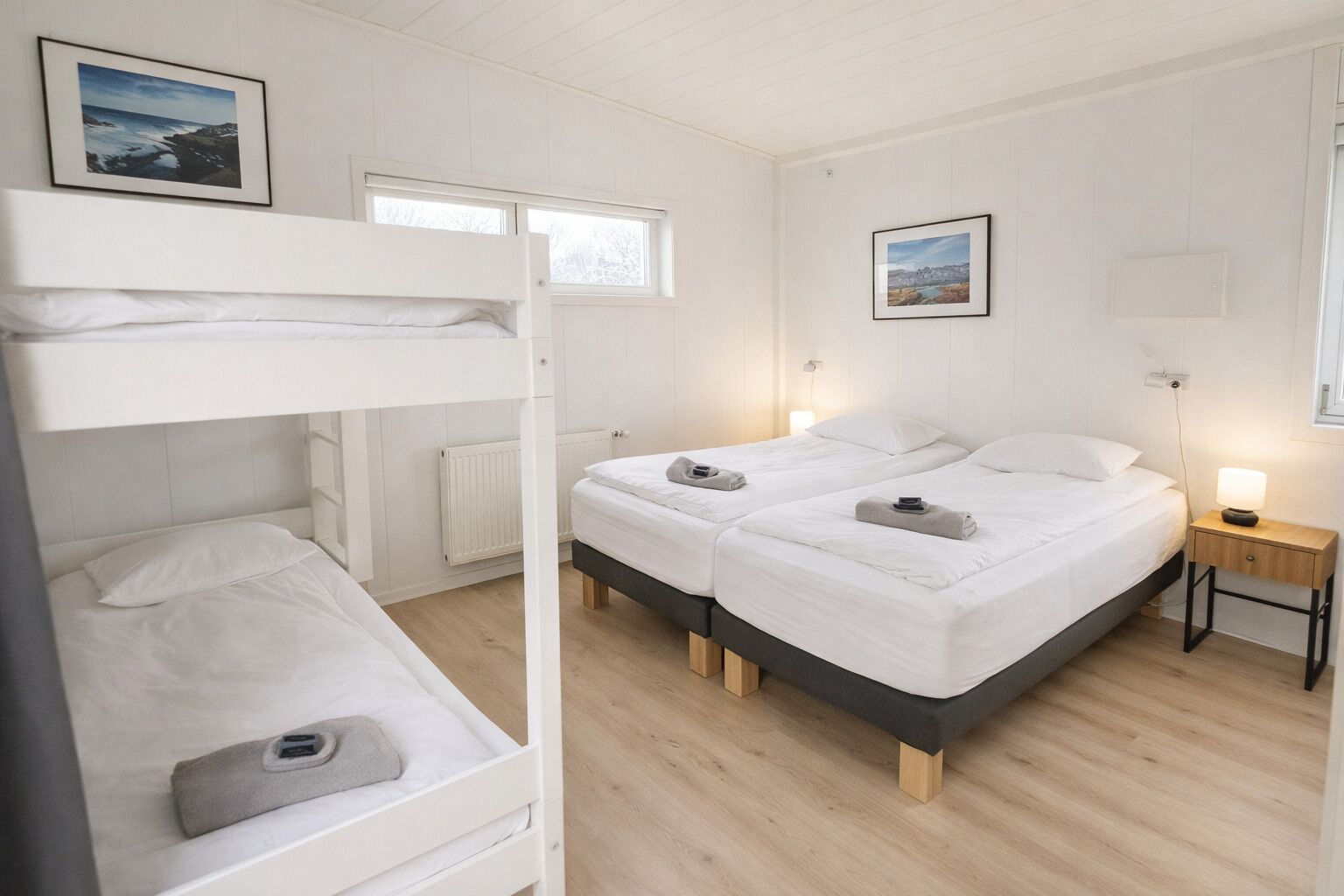 A room with two beds, a bunk bed, a bedside table with a lamp, and two framed pictures on the walls.