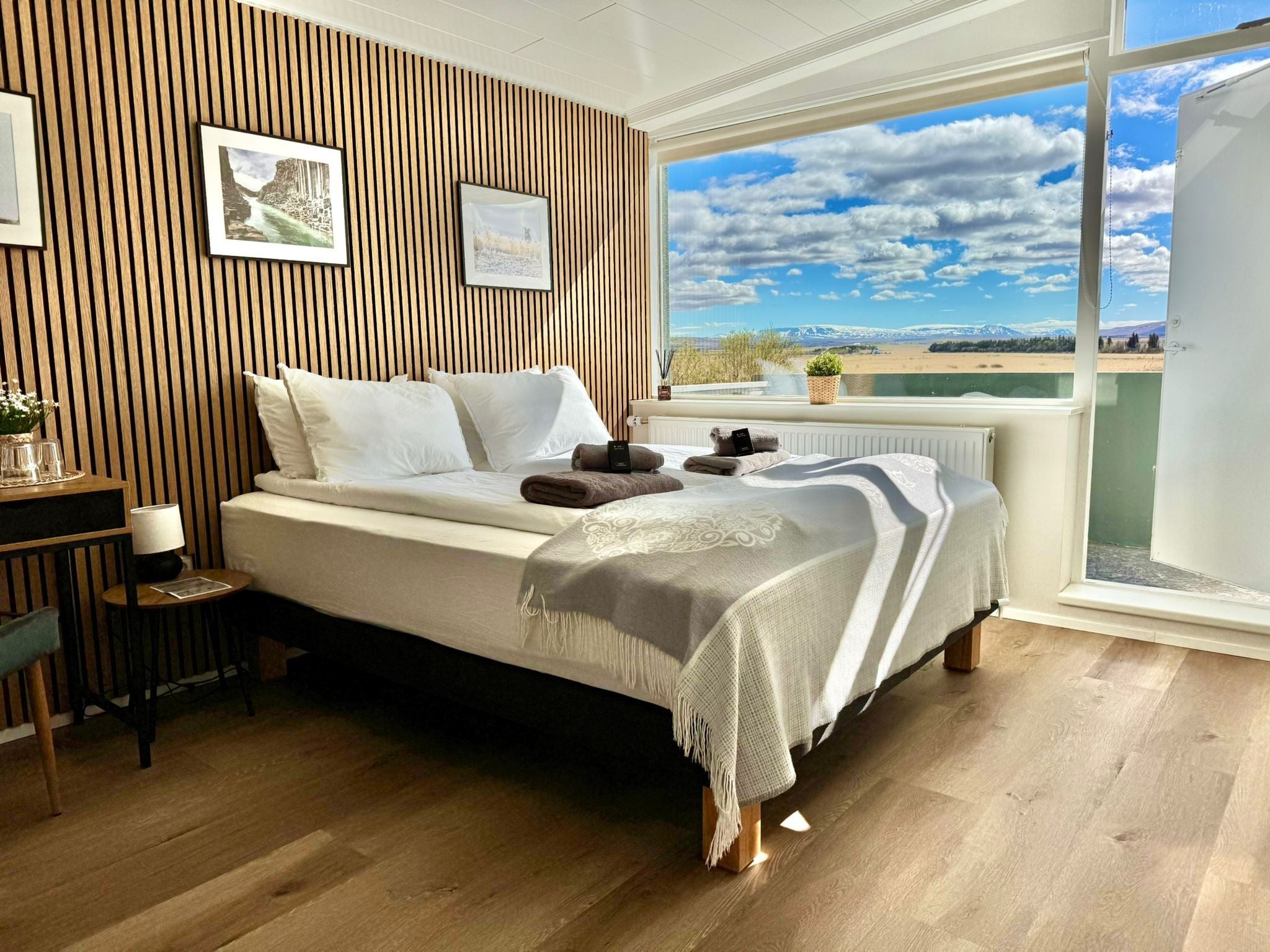 Un dormitorio con cama, pared de listones de madera, cuadros enmarcados, una mesita con vasos y vistas a un paisaje por la ventana.