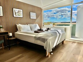 Un dormitorio con cama, pared de listones de madera, cuadros enmarcados, una mesita con vasos y vistas a un paisaje por la ventana.