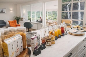 Un bufé de desayuno con diversas opciones de comida y bebidas, incluyendo cereales, leche y miel, en una habitación luminosa con ventanas grandes.