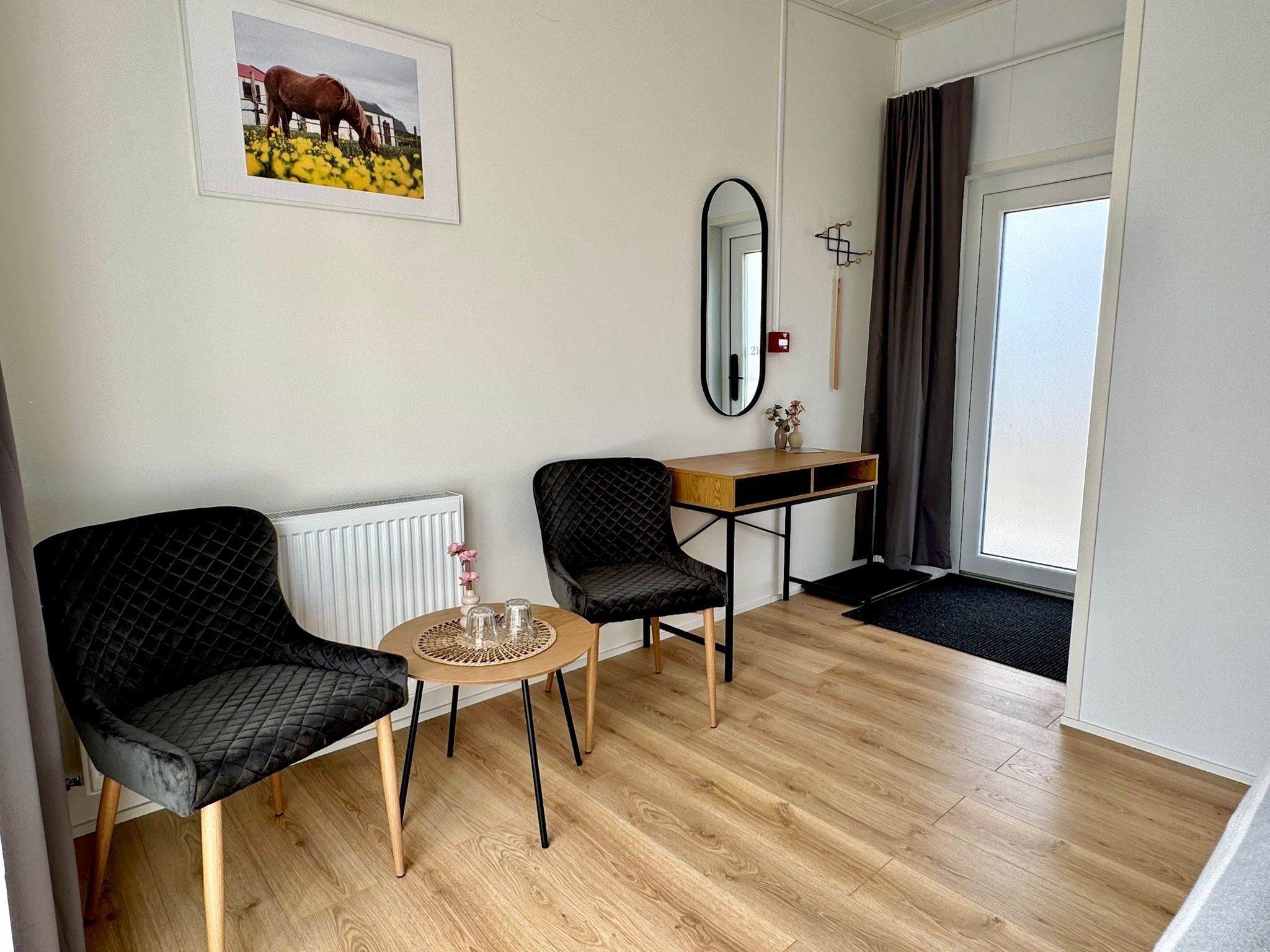 Una habitación moderna con piso de madera, dos sillas negras, una mesa redonda pequeña, un espejo, un radiador, un escritorio de madera y una puerta con cortinas.