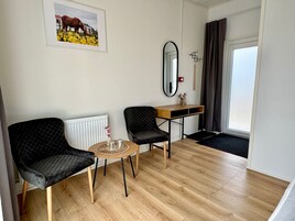 Una habitación moderna con piso de madera, dos sillas negras, una mesa redonda pequeña, un espejo, un radiador, un escritorio de madera y una puerta con cortinas.