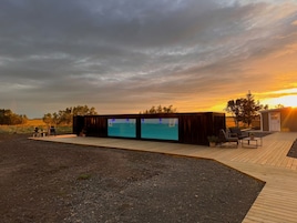 Un espacio exterior moderno con mobiliario, una terraza de madera, dos pantallas grandes y un pequeño edificio con una puerta.