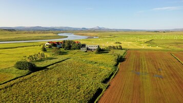Un paisaje rural con una casa grande rodeada de campos, un río y montañas a lo lejos.
