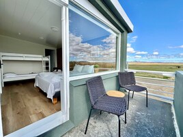 Un dormitorio con una litera y una cama individual, un balcón con dos sillas y una mesita, y un amplio ventanal con vistas al paisaje.