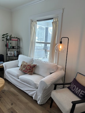 Un canapé blanc agrémenté d’un coussin à motifs floraux, une table d’appoint, une lampe sur pied et une bibliothèque contenant des livres et une plante.