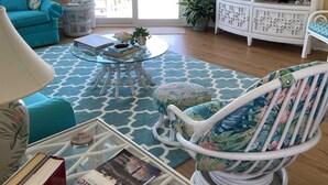 Una sala de estar con un sofá color turquesa, un sillón blanco, una mesa de centro de vidrio y un televisor sobre un mueble blanco.