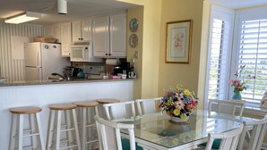 Una cocina con gabinetes blancos, un microondas y un refrigerador. Hay una zona de comedor con una mesa de vidrio y sillas blancas con cojines azul turquesa.