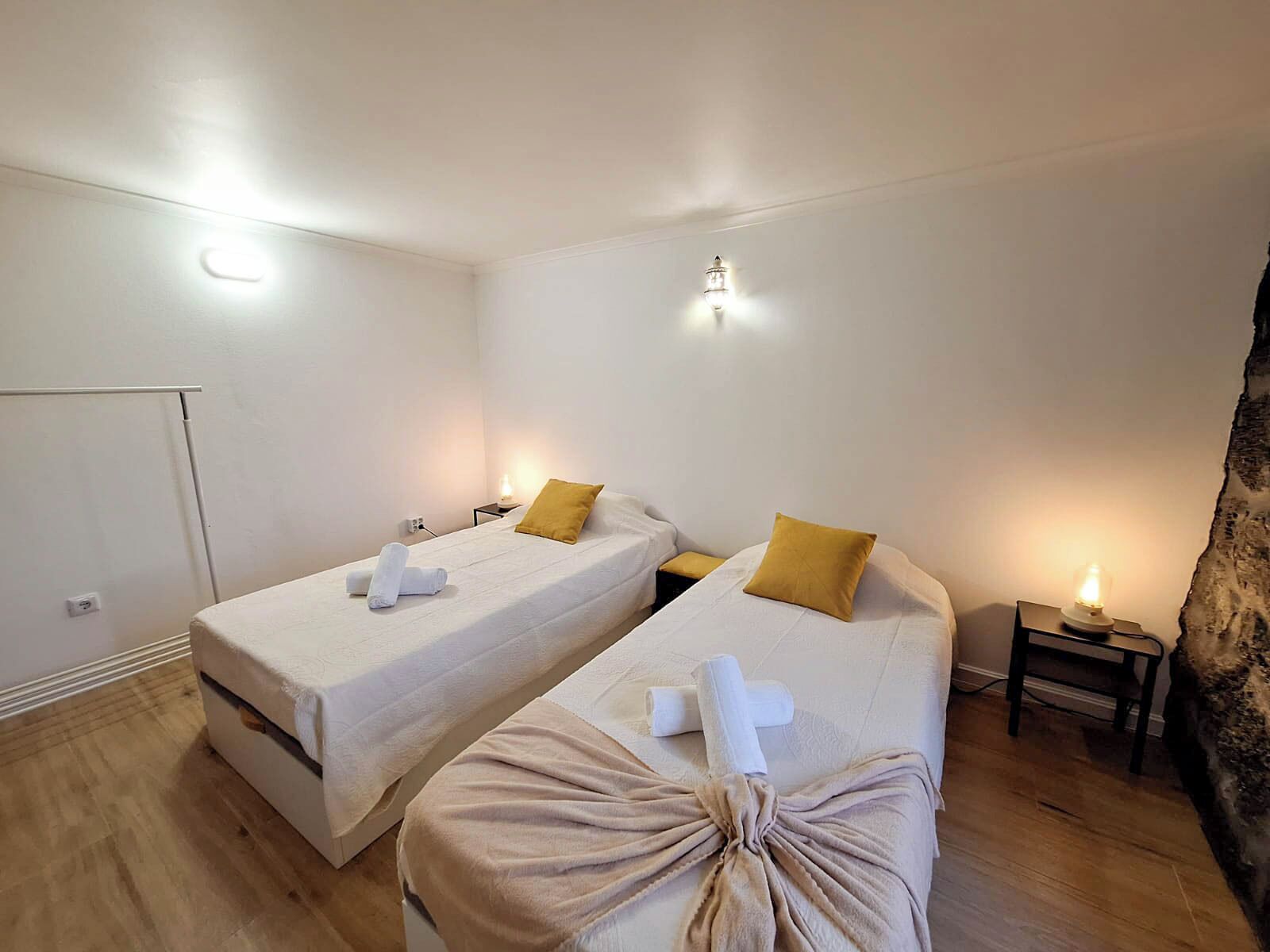 Une chambre avec deux lits, chacun recouvert d’une courtepointe blanche et agrémenté d’un oreiller jaune. On y trouve des tables de chevet avec des lampes, et le sol est recouvert de parquet.