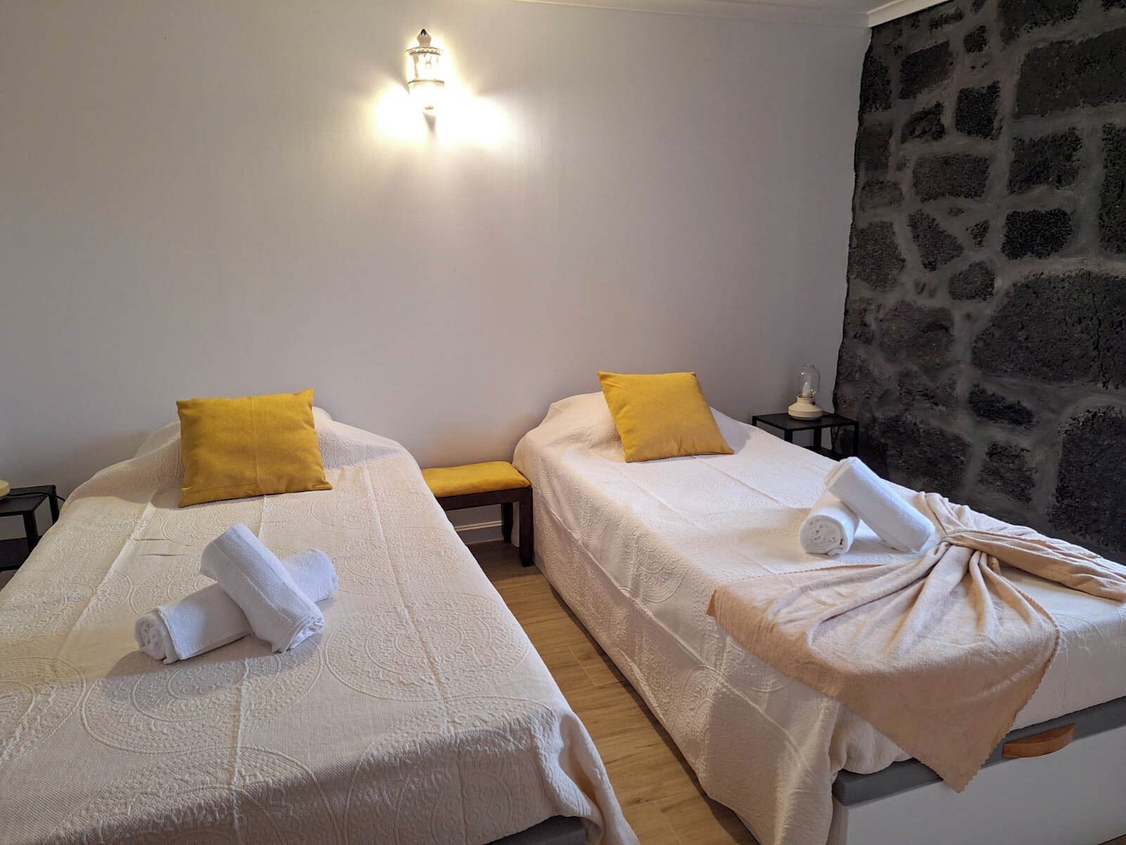 Dos camas individuales con ropa de cama blanca y almohadas amarillas, un banco y una pared de piedra.