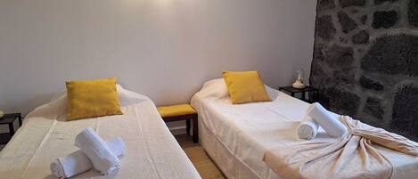 Dos camas individuales con ropa de cama blanca y almohadas amarillas, un banco y una pared de piedra.