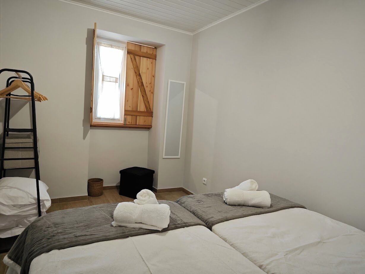A hotel room with two beds, a wooden wardrobe, a window with curtains, a ladder, and a trash bin.