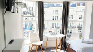A hotel room with a bed, a desk, a chair, a small table, a trash bin, and a large window with curtains. - Central Wave Brighton  (Brighton)