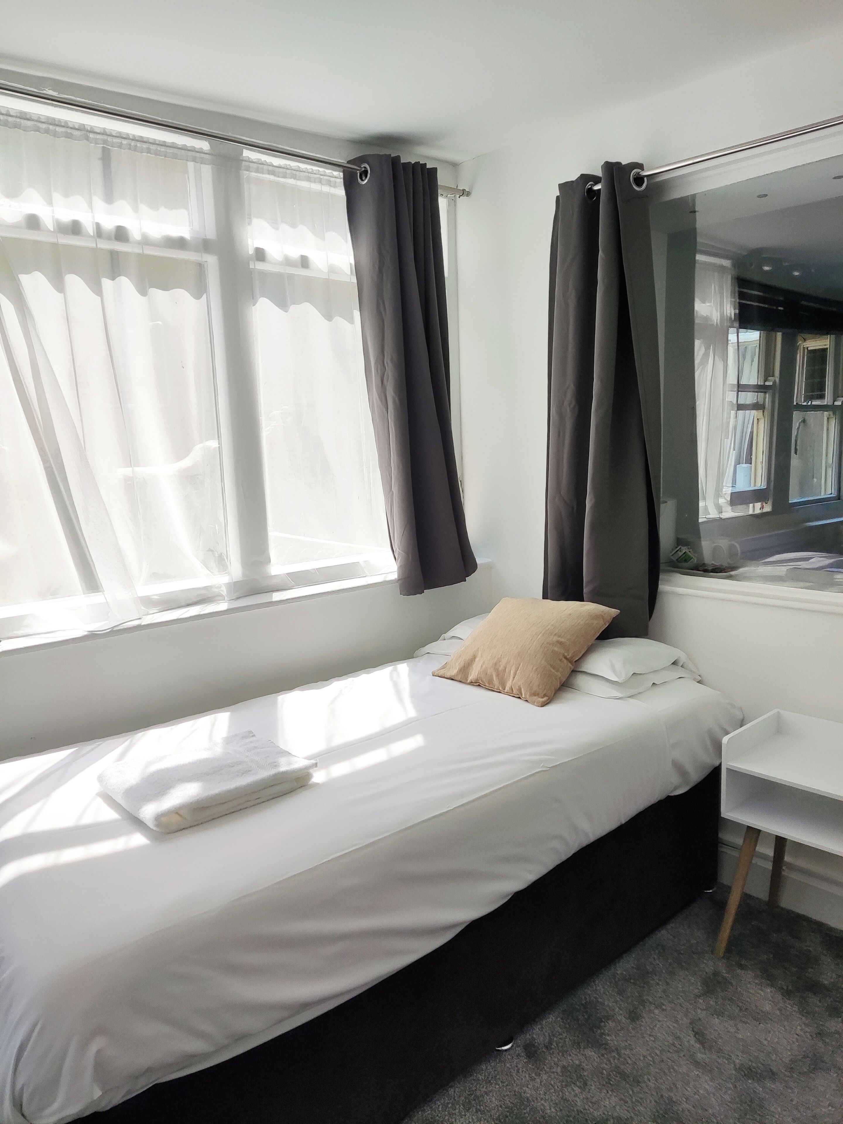 Een eenpersoonsbed met een wit matras en een beigekleurig kussen, een klein nachtkastje en een raam met gordijnen.