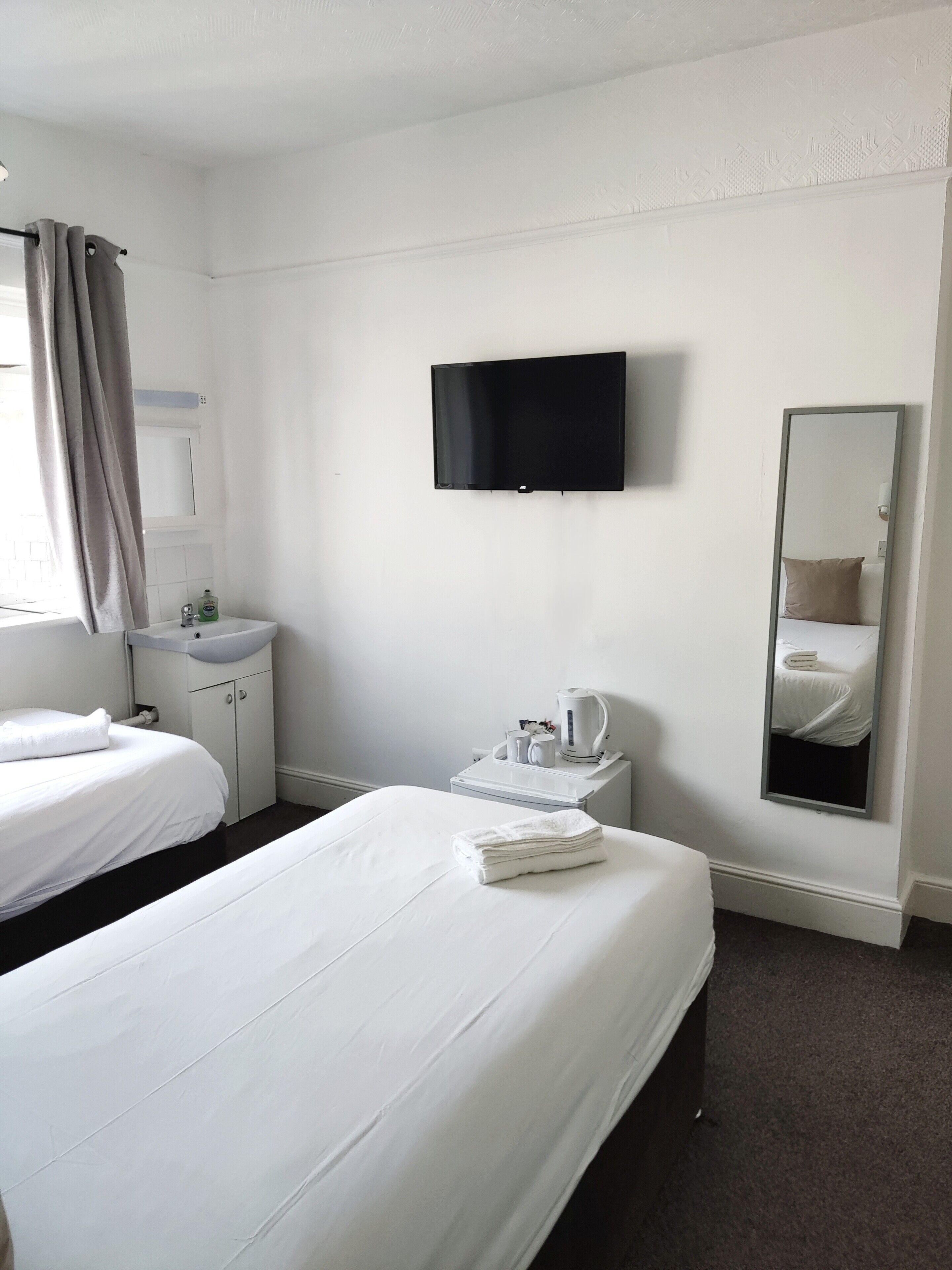 A hotel room with two beds, a flat-screen TV, a mirror, a bedside table with a kettle, and a window with curtains.