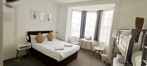 Ein Hotelzimmer mit einem Bett, Nachttischen, einem Schreibtisch, Stühlen, einem Fenster mit Vorhängen und einem Stockbett.
