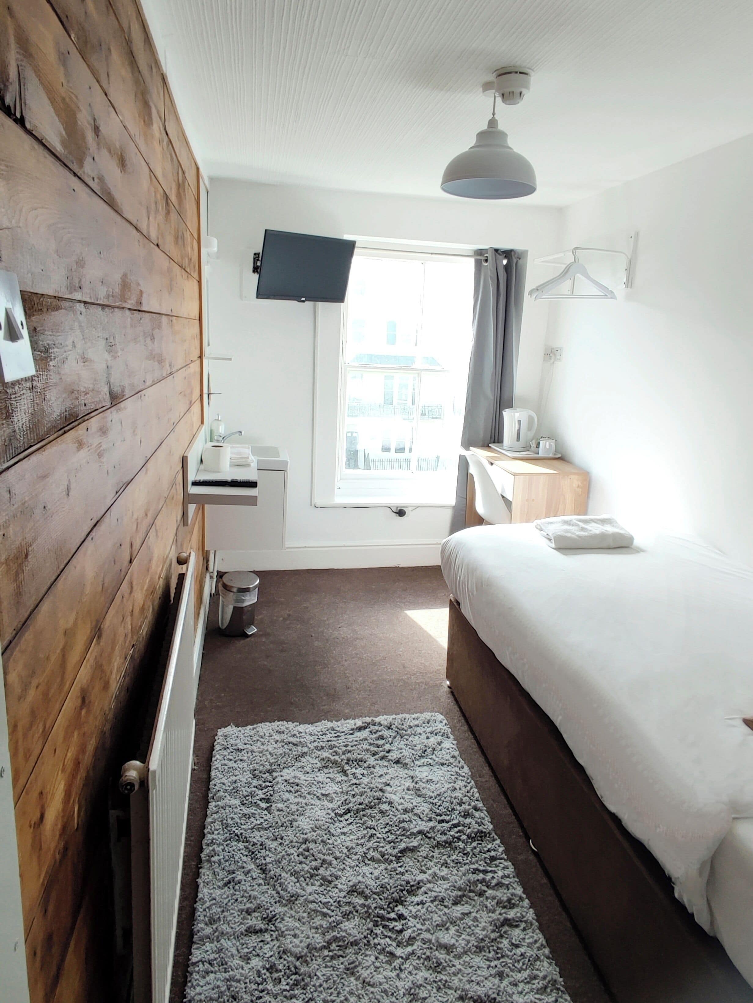 A bedroom with a wooden wall, a bed, a desk, a window, and a TV.