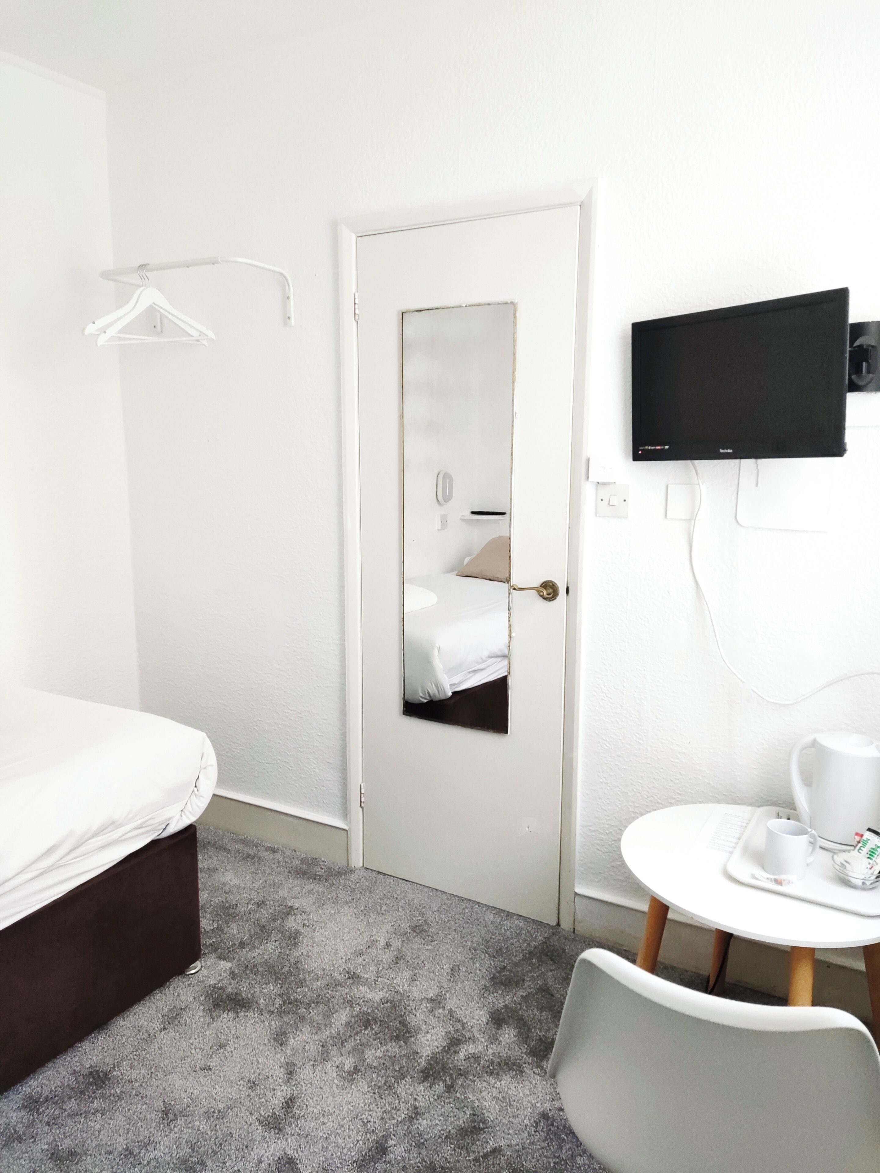 Habitación de hotel con cama, una mesita con cafetera, televisor montado en la pared y una puerta con espejo.