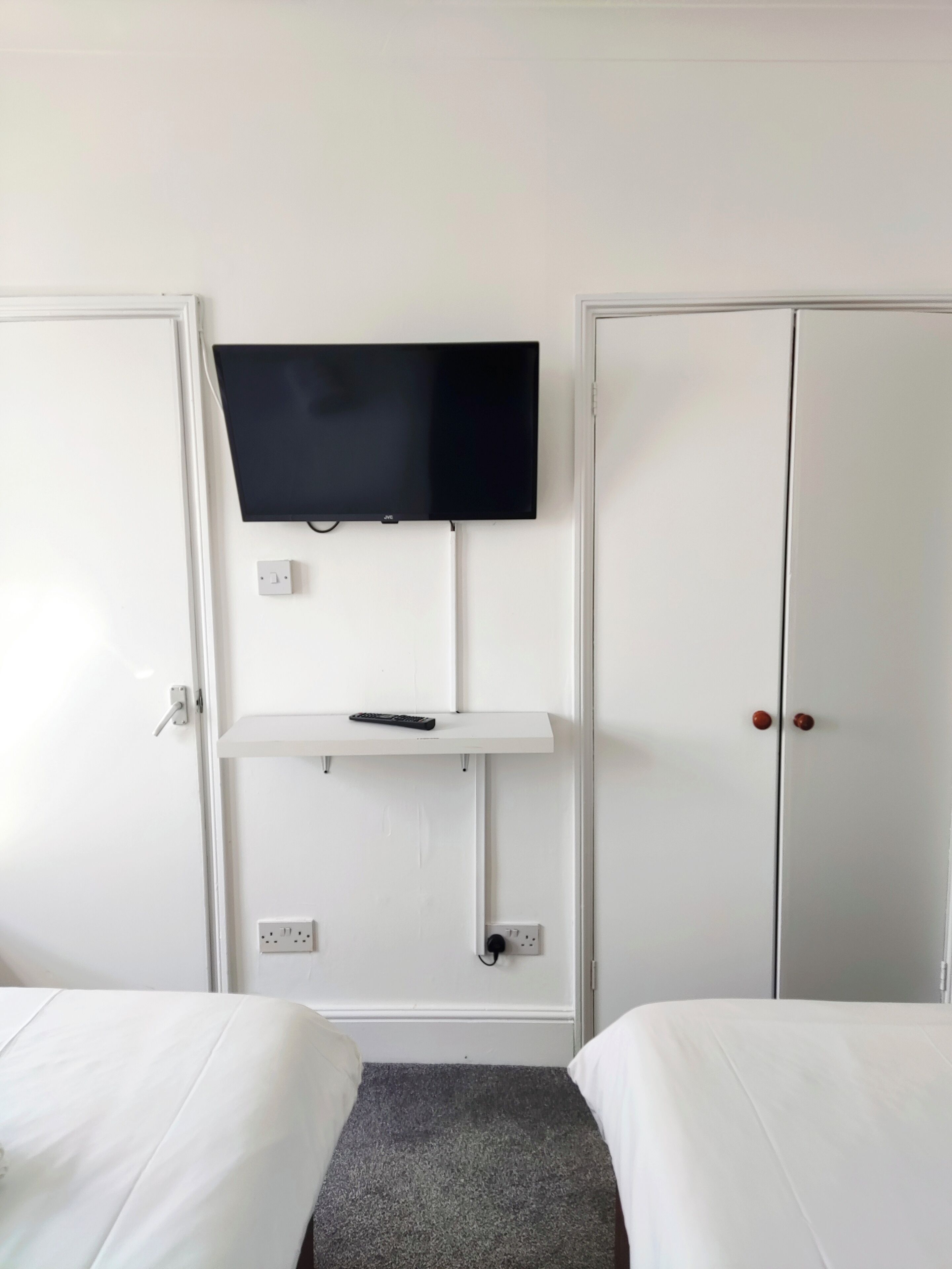 Une chambre d’hôtel avec un téléviseur fixé au mur, une étagère avec une télécommande, deux armoires et un lit recouvert d’une moquette grise.
