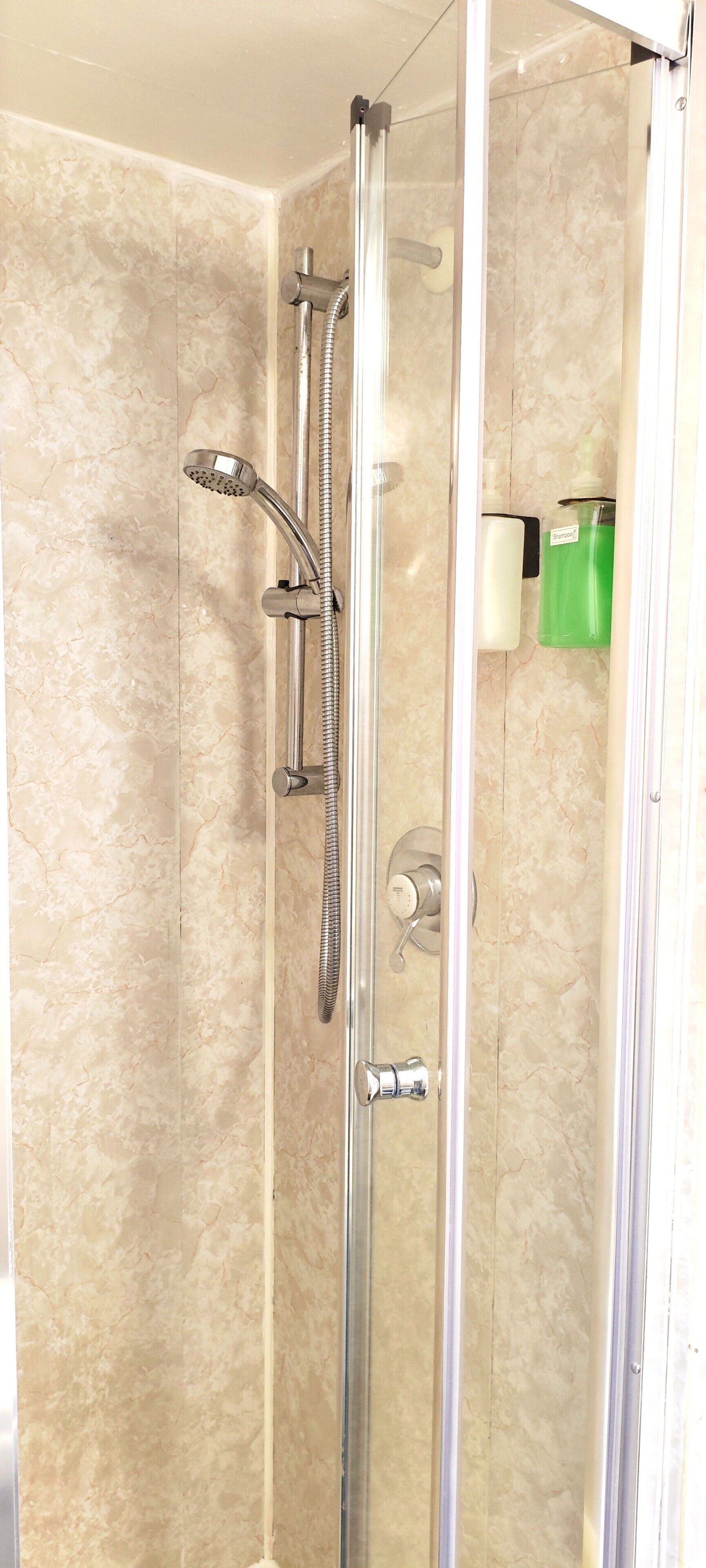 Une douche fermée par des parois en verre, dotée d’un pommeau de douche à main, d’un robinet mural et d’un distributeur de savon.