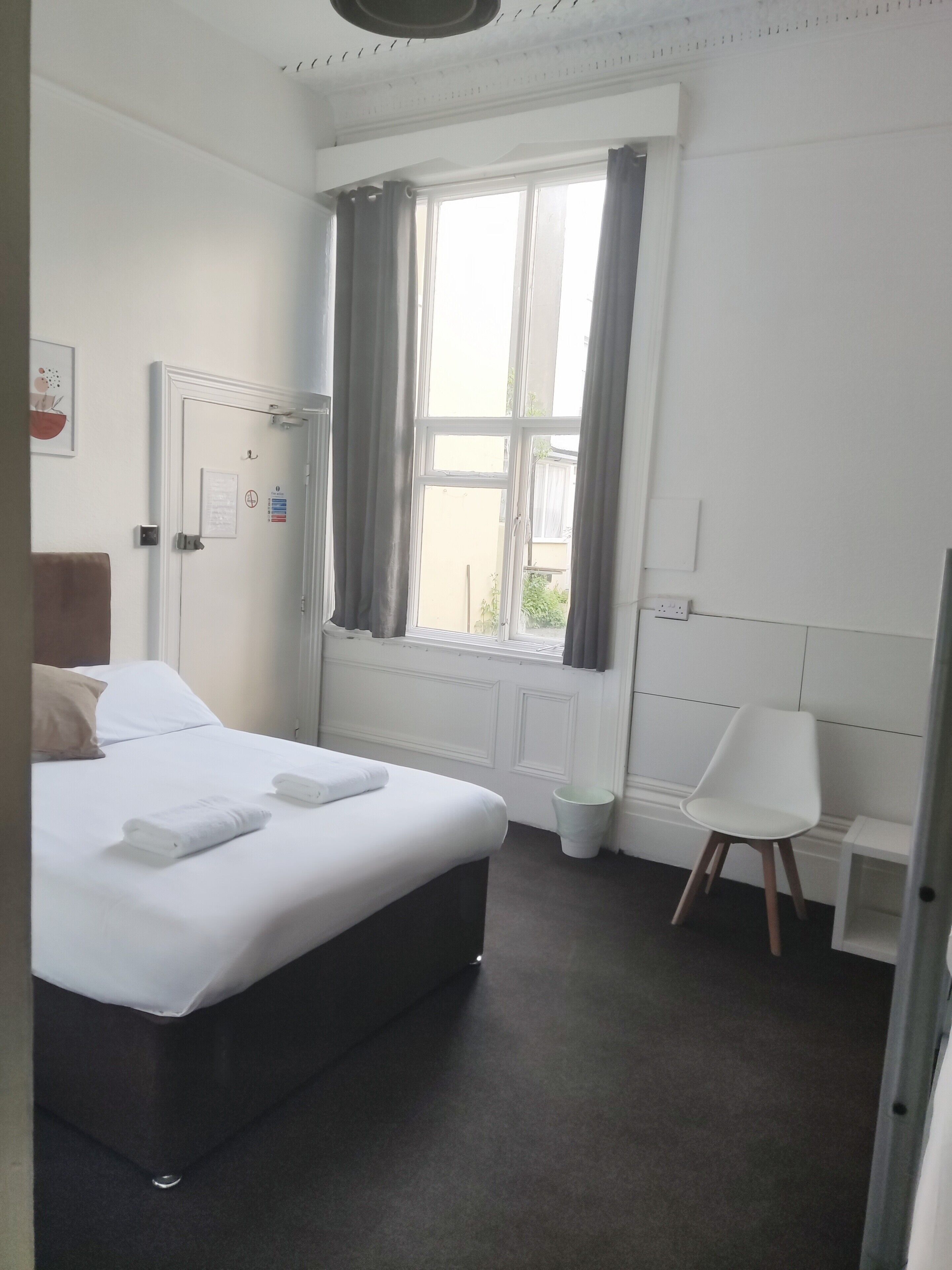 A hotel room with a bed, a chair, a window with curtains, a door with a sign, and a wall-mounted light fixture.