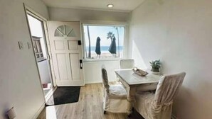 A room with a wooden floor, a white door, a window with a view of the beach, a table with a plant, and a chair. - Manhattan Beach Beachfront Charming On The Strand (Manhattan Beach)